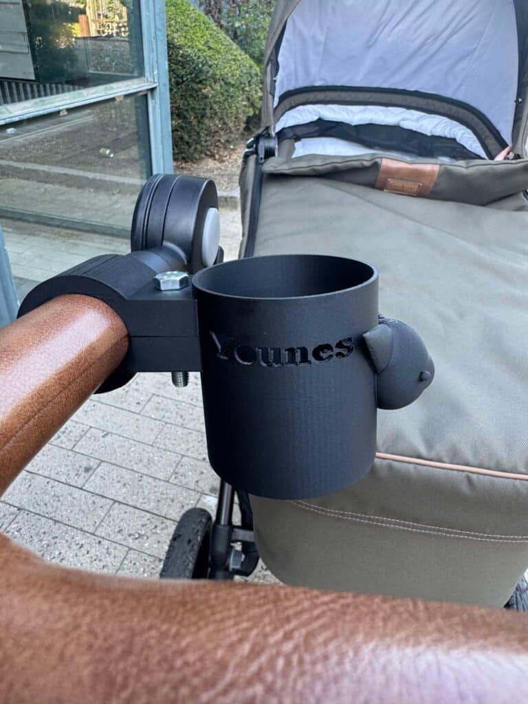 A black 3D-printed cup holder is attached to a stroller handle, with the name 