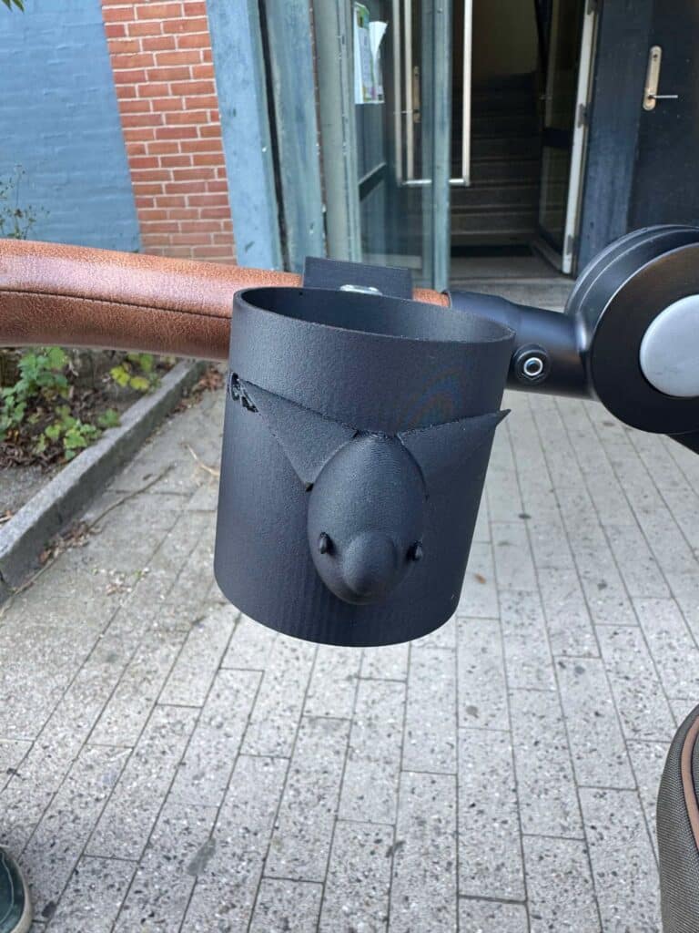 A close-up shows a black 3D-printed stroller cup holder with a fox head decoration mounted to a brown rounded handlebar with a bolt.