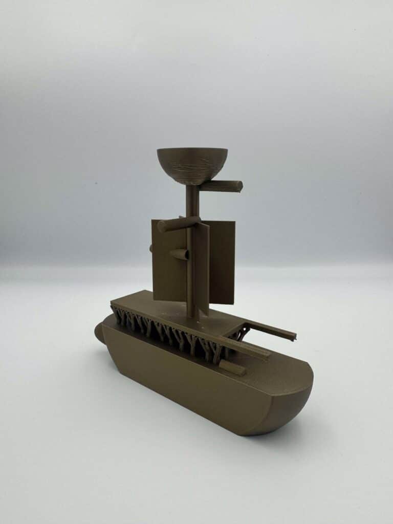 A metallic gold 3D-printed boat-like model has a rounded hull, a central mast and a bowl-shaped element at the top, with geometric supports and small printed details visible.