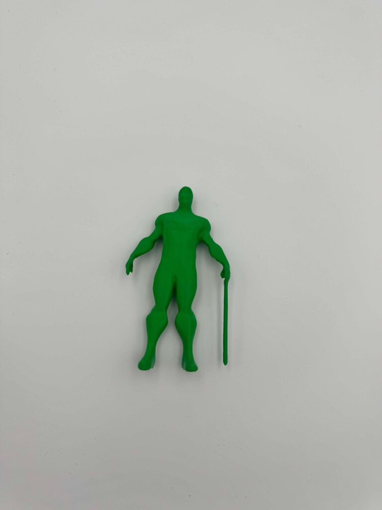 A green 3D-printed human figure is standing upright and holding a long cane. The model has a simple abstract shape with smooth surfaces and minimal facial detail.