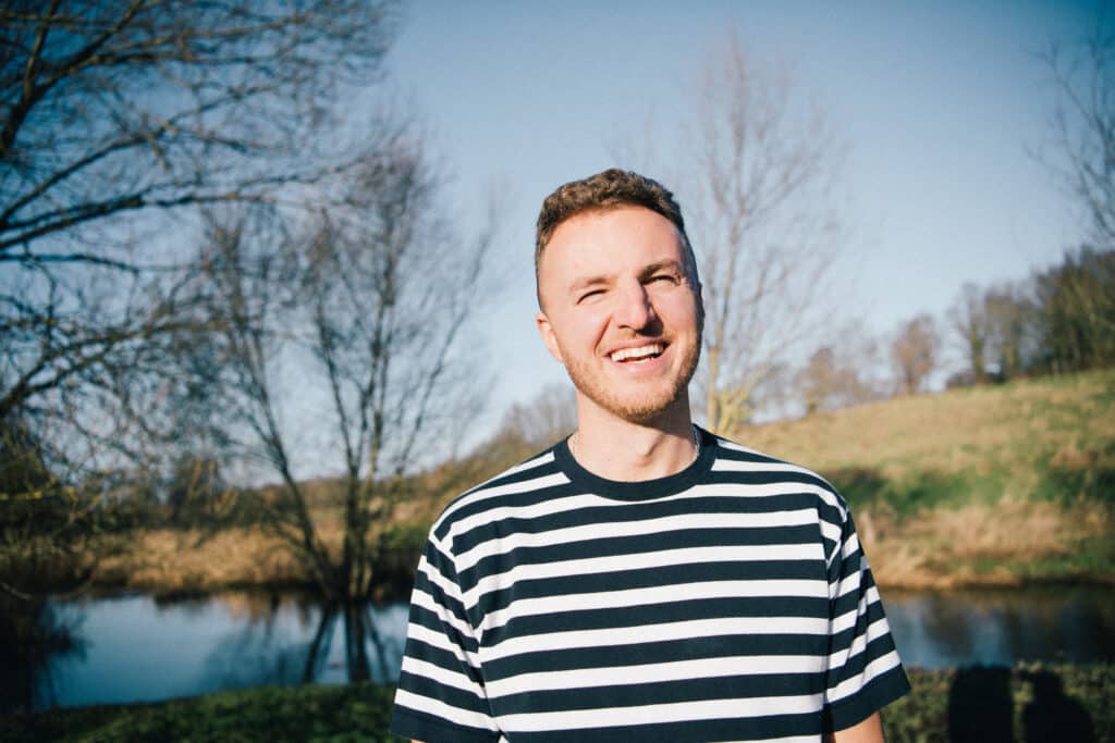 Edis is standing outdoors and smiling in sunlight, wearing a striped shirt with trees, water and grassy hills behind him.