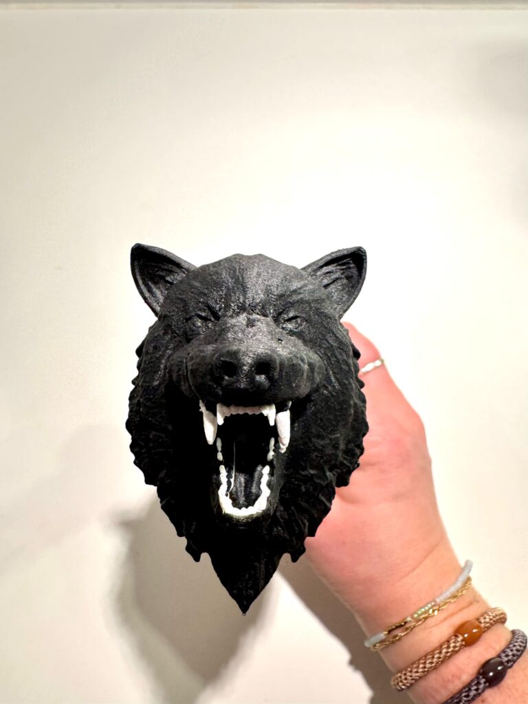 A close-up of a black wolf-shaped knife pommel shows textured fur, an open mouth and white fangs, with the handle held in a hand wearing bracelets.