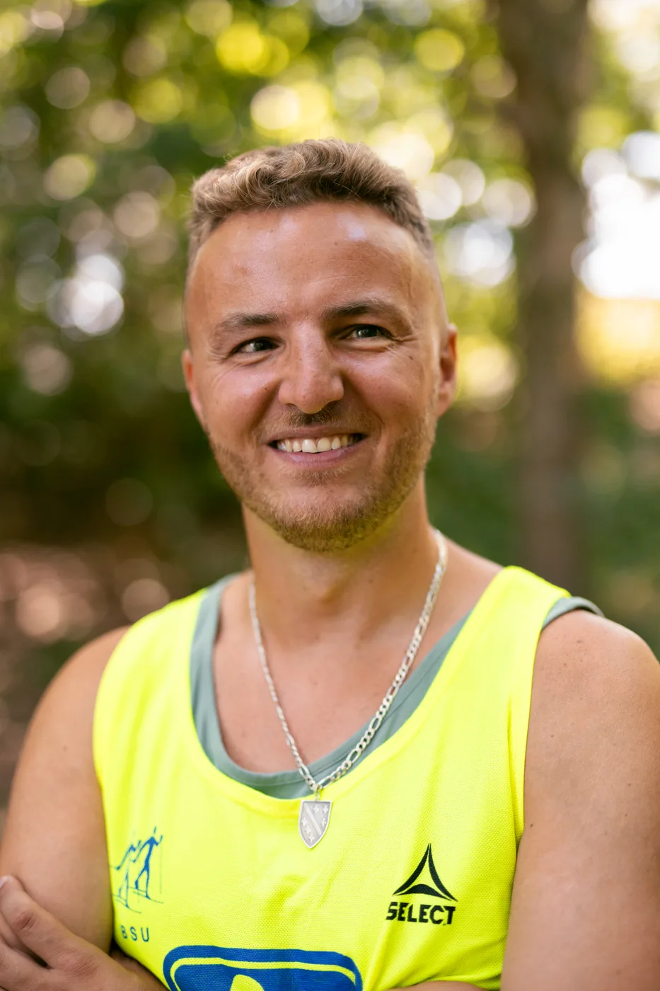A close portrait of Edis smiling in woodland, with his arms folded. He is wearing a bright yellow high-visibility vest and a silver chain, with soft green trees behind him.