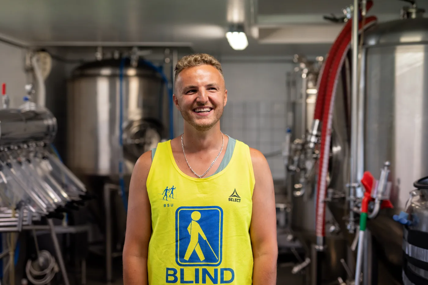 Edis is standing and smiling inside a brewery, surrounded by polished stainless-steel brewing tanks and a coiled red hose. He is wearing a bright yellow high-visibility vest printed with the word "BLIND" and the blue-and-white symbol of a person walking with a long cane.