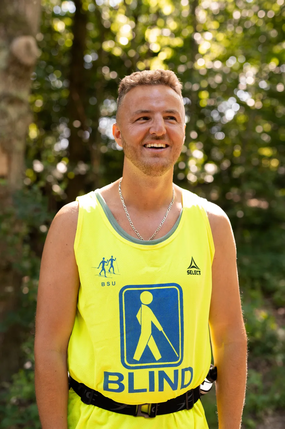 Edis Adilovic, the blind founder of Accessible3D, smiles outdoors in woodland sunlight. He wears a bright yellow high-visibility vest printed with the word “BLIND” and the blue-and-white international symbol of a person walking with a long cane.