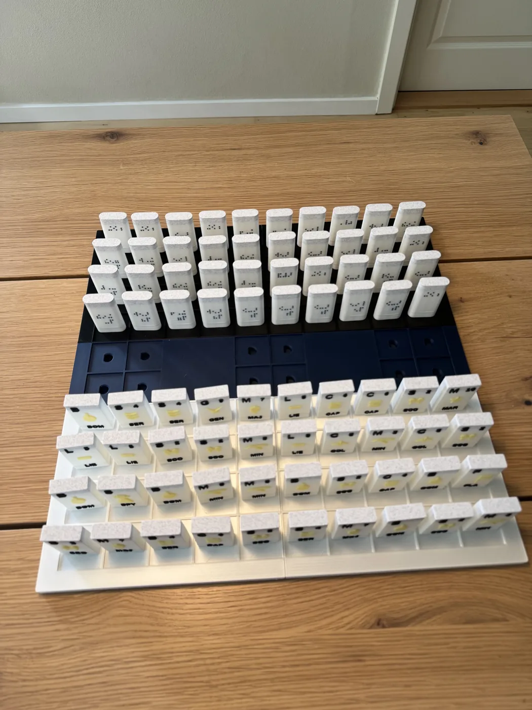 A 3D-printed tactile Stratego board sits on a wooden floor, with a navy and white grid and two armies of upright playing pieces marked with Braille dots and short raised letters.