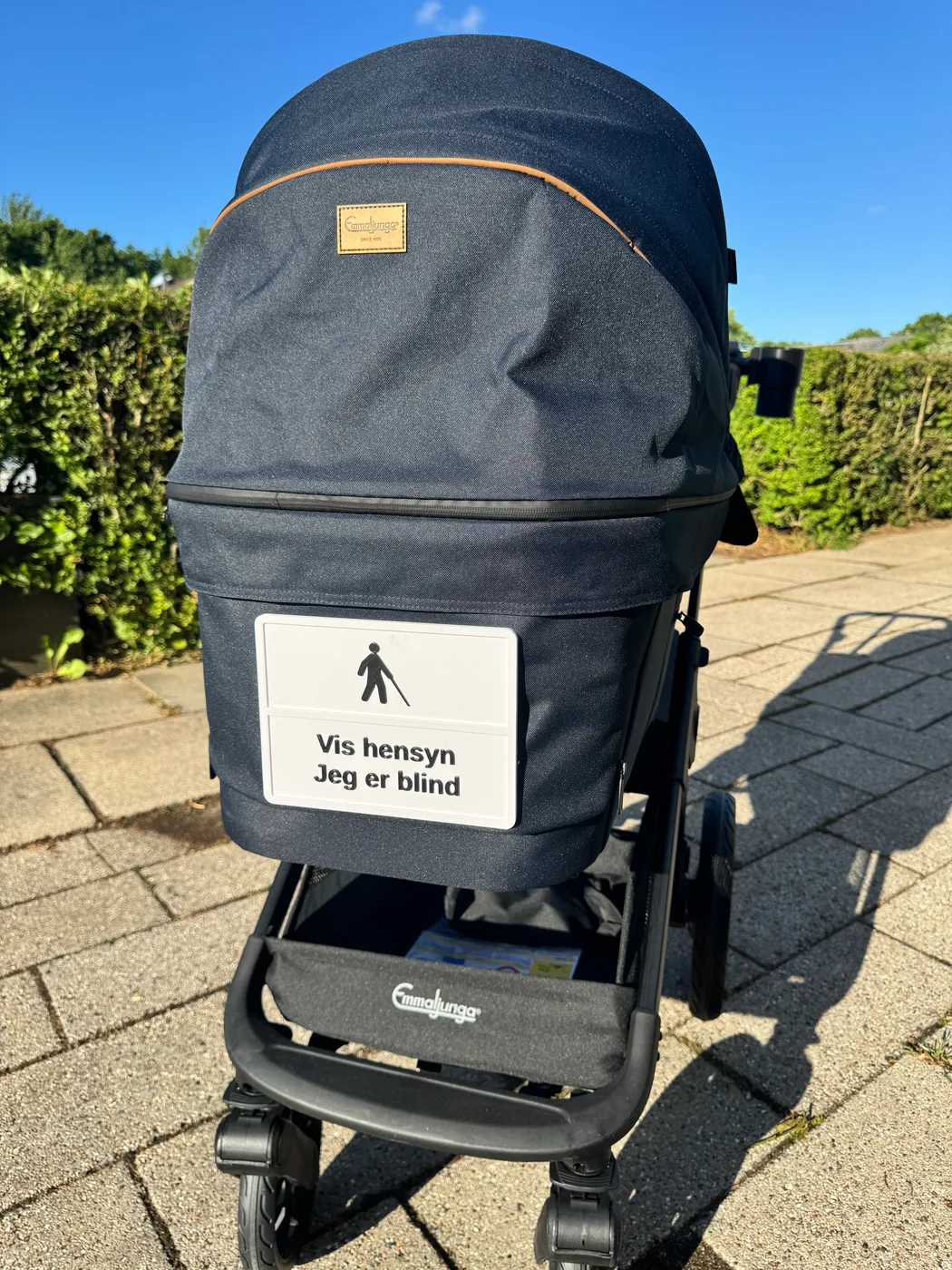 A parametric 3D-printed blind awareness sign is mounted on a baby stroller hood outdoors. It shows the text Vis hensyn Jeg er blind and the ISO 7001 white-cane symbol.