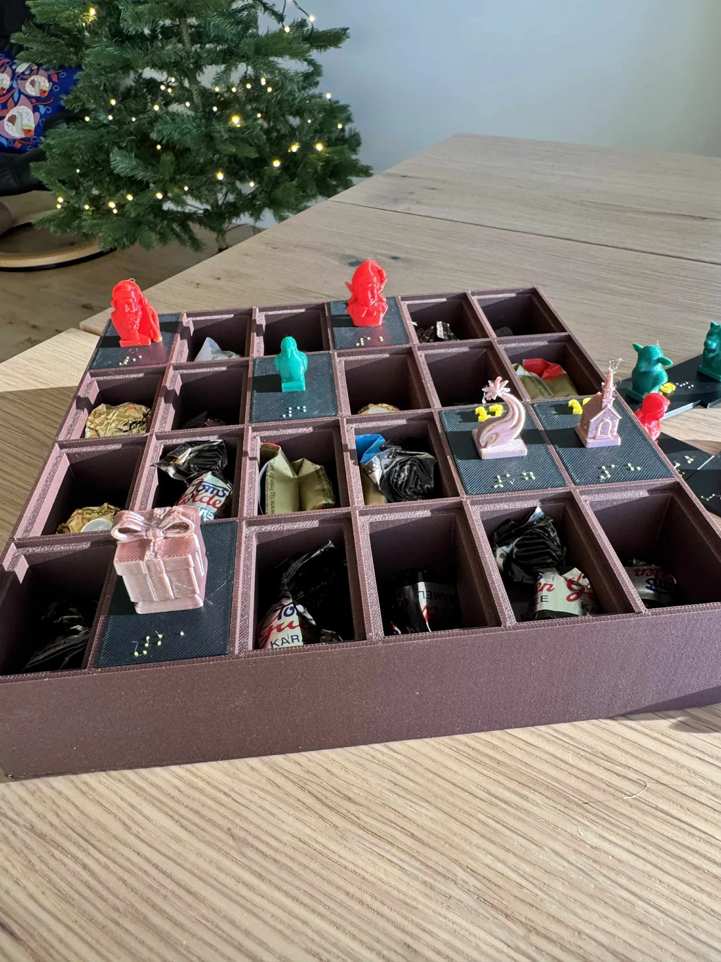 A top-down view shows a 3D-printed Braille Advent calendar on a wooden table, with 24 small compartments, raised Braille numerals, colourful ornament busts and foil-wrapped chocolates.