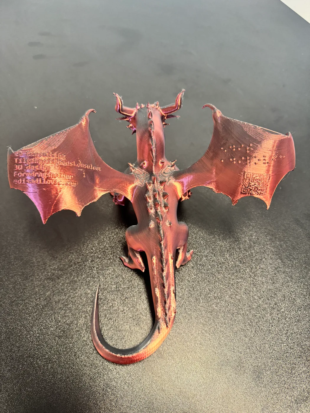 A top-down view shows a 3D-printed dragon name plate in an iridescent copper-to-purple finish, with spread wings, raised lettering, a ridged spine, clawed legs and a curling tail.