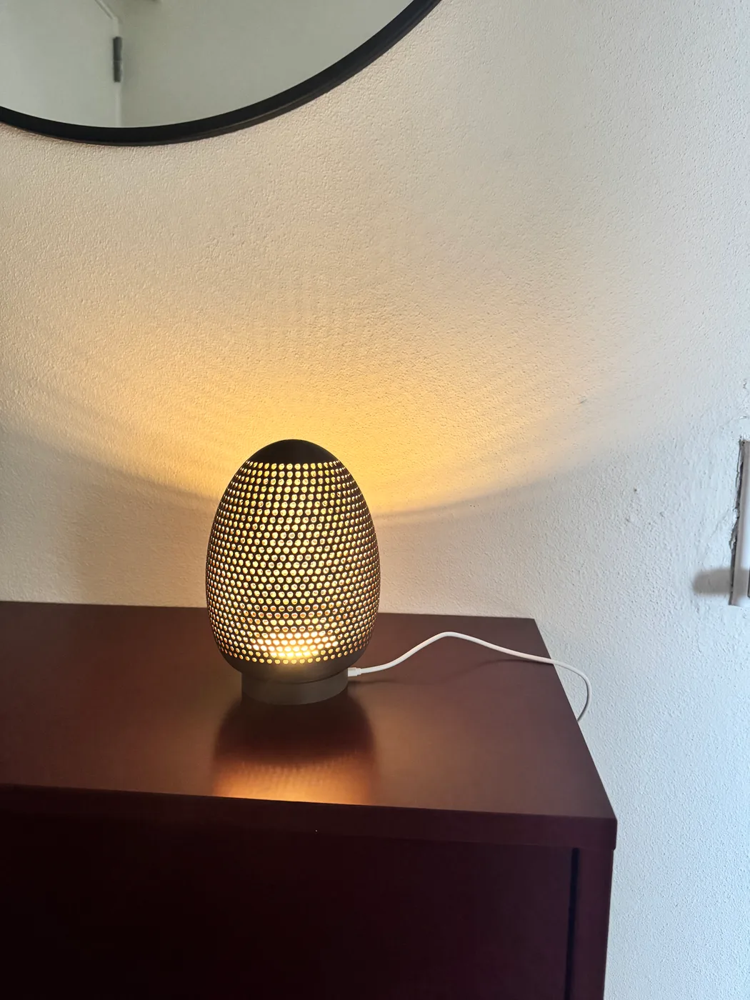 A 3D-printed egg lamp sits on a wooden table, with a warm glow shining through its perforated Voronoi-patterned shell.