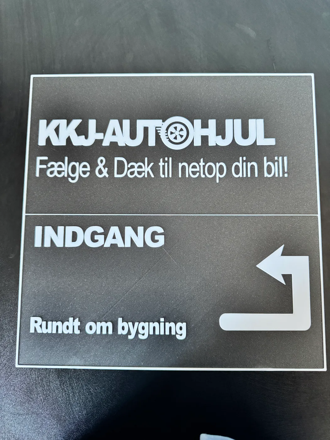 A large 3D-printed workshop directional sign shows high-contrast white raised text on a dark textured background, displaying the brand KKJ-AUTOHJUL.