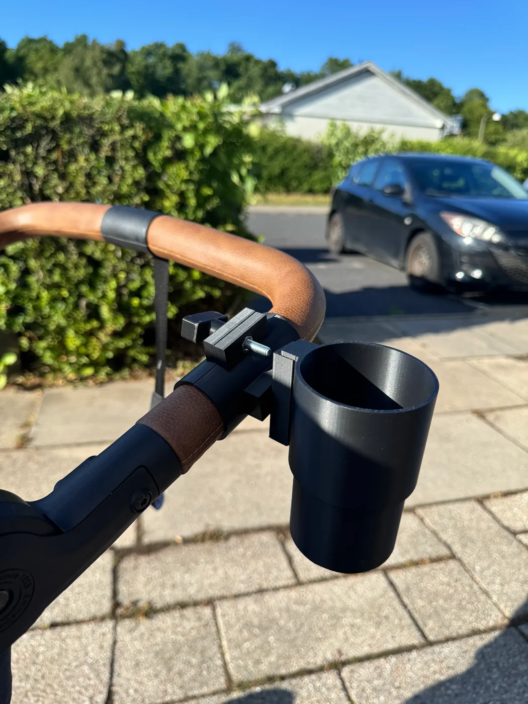 A finished black 3D-printed baby stroller cup holder is mounted outdoors on a pram handlebar with an adjustable clamp.
