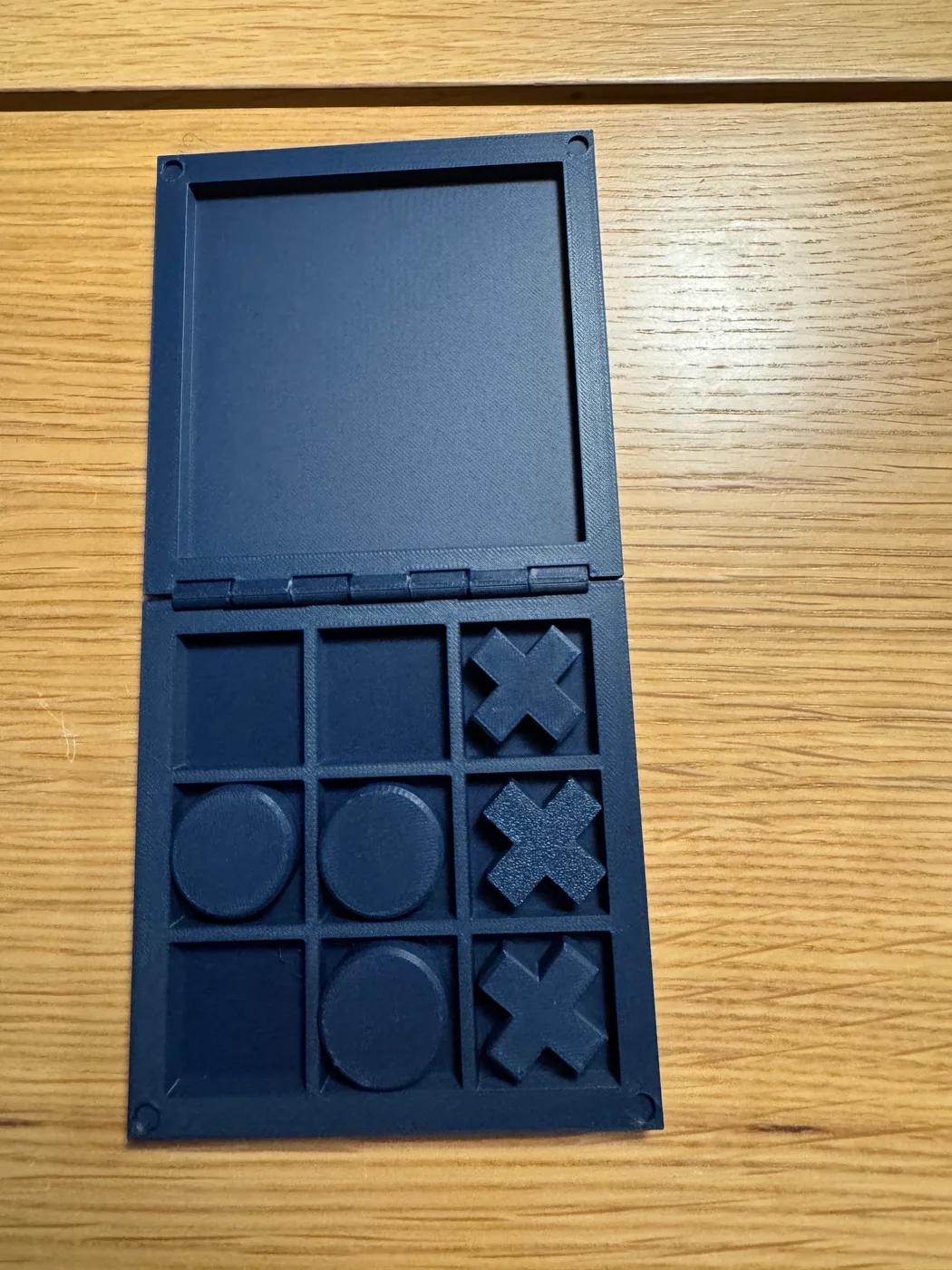 An accessible 3D-printed tactile tic-tac-toe travel set is open, showing deep grid pockets and distinct cross and circle pieces.